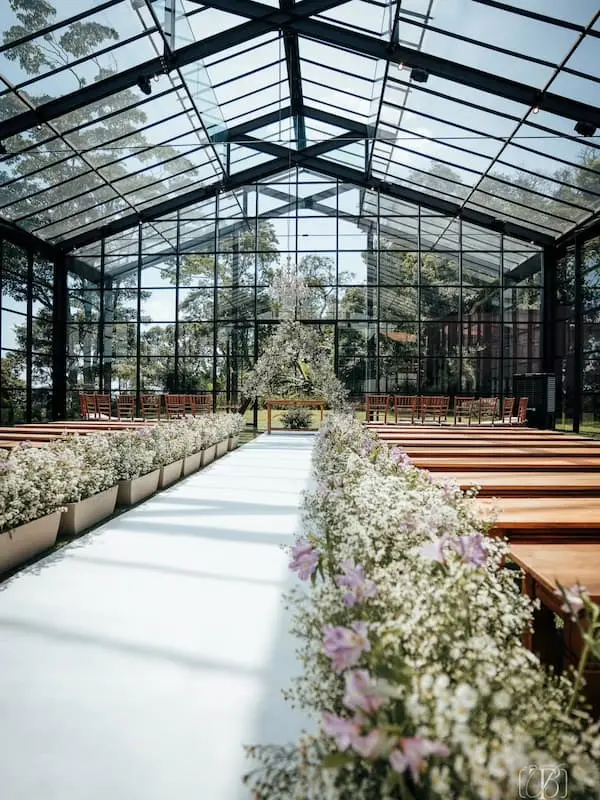 Casa de vidro para cerimônia de casamento no Mirante Juruá, com vista para a natureza, bancos de madeira e caminho de flores brancas e lilases. O ambiente interno garante proteção climática com iluminação natural, ilustrando a importância de um plano B elegante ao decidir qual o melhor mês para casar.