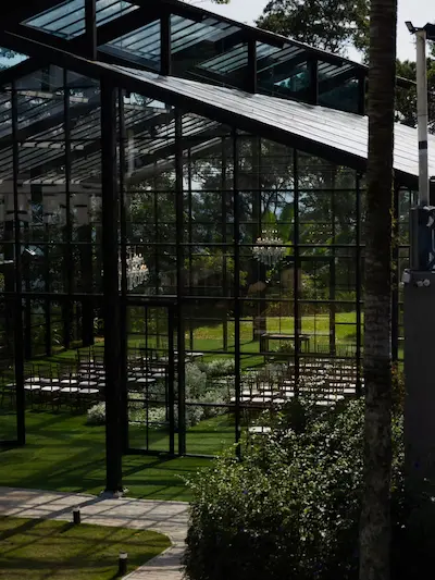 Interior da Casa de Vidro montada para cerimônia. O espaço possui cadeiras escuras alinhadas sobre um gramado sintético verde, arranjos rasteiros de flores brancas e lustres de cristal pendurados no teto. As paredes de vidro revelam a vegetação e a luz natural do lado de fora.