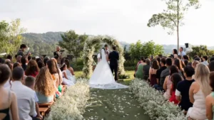 Visão panorâmica de uma cerimônia de casamento ao ar livre no gramado do Mirante Juruá, capturando a essência de um casamento no inverno. O casal de noivos, visto de costas, está em pé sob um grande arco circular decorado com flores brancas e folhagem, no altar. Uma longa passarela central, ladeada por densas bordas de flores brancas (mosquitinho) e pétalas no tapete verde, leva os convidados, sentados em cadeiras de madeira, até os noivos. Atrás do altar, estende-se uma vista deslumbrante da serra e paisagens arborizadas da Serra da Cantareira sob um céu limpo e azul claro. Fotógrafos e convidados assistem à celebração em um ambiente natural e rústico-chique.