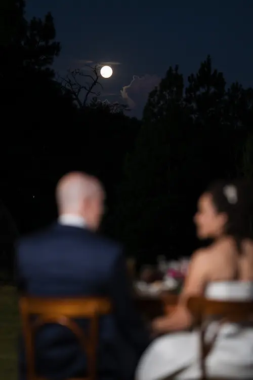 Noivos à mesa durante a festa, sob a lua cheia, celebrando um casamento exclusivo ao ar livre.