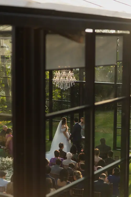 Cerimônia de casamento exclusivo vista através das vidraças da Casa de Vidro, com lustre de cristal e noivos no altar.