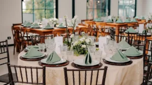 Mesas redondas preparadas para o jantar no salão principal do Mirante Juruá, decoradas com toalhas claras, arranjos florais brancos e verdes no centro, louças, taças de vidro e guardanapos verdes elegantemente dobrados sobre os pratos. Ao fundo, grandes janelas de vidro deixam a luz natural entrar. A imagem ilustra o cuidado com os detalhes que garantem o conforto de cada convidado de casamento.