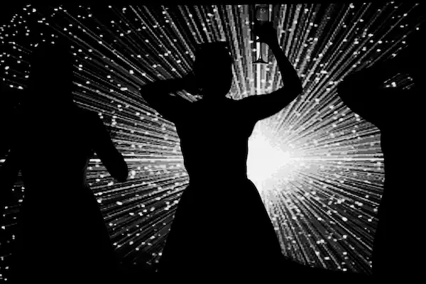 Fotografia em preto e branco (silhueta) de três pessoas se divertindo intensamente na pista de dança. A pessoa no centro ergue uma taça para o alto com uma mão e toca a cabeça com a outra, enquanto dança de costas para a câmera em frente a um painel de LED que projeta linhas luminosas convergentes (efeito "starburst"). A imagem captura a energia alta que transforma a festa em uma experiência imersiva para o convidado de casamento.